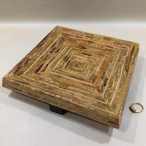 May include: A square wooden tray with a geometric pattern of concentric squares. The tray is made of light brown wood with darker brown and tan accents. A small gold ring with small stones sits on the side. The tray is supported by a small black block.