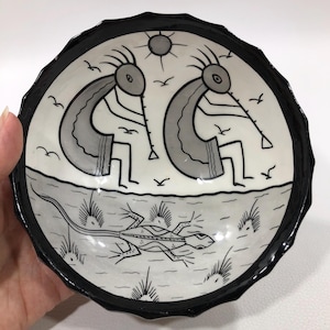 May include: A round ceramic bowl with a black scalloped rim. The bowl features a black and white design of two Kokopelli figures playing flutes, birds, a sun, and a lizard.
