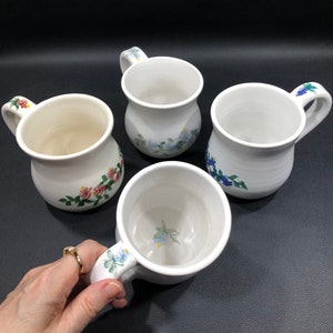 May include: Four white ceramic mugs with floral designs. The mugs have a rounded shape and a handle. The designs include blue, pink, and green flowers.