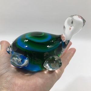 May include: A clear glass turtle figurine with a vibrant shell design. The shell features concentric circles of blue and green. The turtle's head and flippers are transparent, and the figurine is held in a hand.