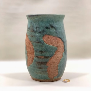 May include: A cylindrical ceramic vase featuring a textured surface with a mottled teal and blue glaze that partially reveals the raw, unglazed brown clay body underneath. A small gold ring with clear stones sits on the surface next to the vase.