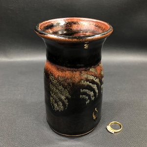 May include: A handcrafted ceramic vase with a dark brown and black glaze. The vase has a flared rim and a cylindrical body with a textured, organic pattern in shades of brown and green. A gold ring with small stones sits next to the vase.