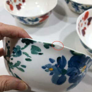 May include: White ceramic bowls with floral designs in shades of blue, red, and green. The bowls have a smooth, glossy finish and a slightly irregular rim. The image shows a set of bowls, with one bowl being held.
