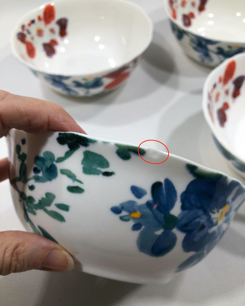 May include: White ceramic bowls with floral designs in shades of blue, red, and green. The bowls have a smooth, glossy finish and a slightly irregular rim. The image shows a set of bowls, with one bowl being held.