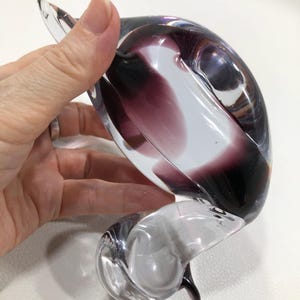 May include: A clear glass sculpture with a swirling design, featuring shades of purple and white. The art glass piece has a curved, organic shape and is held by a hand. The sculpture is a decorative item.