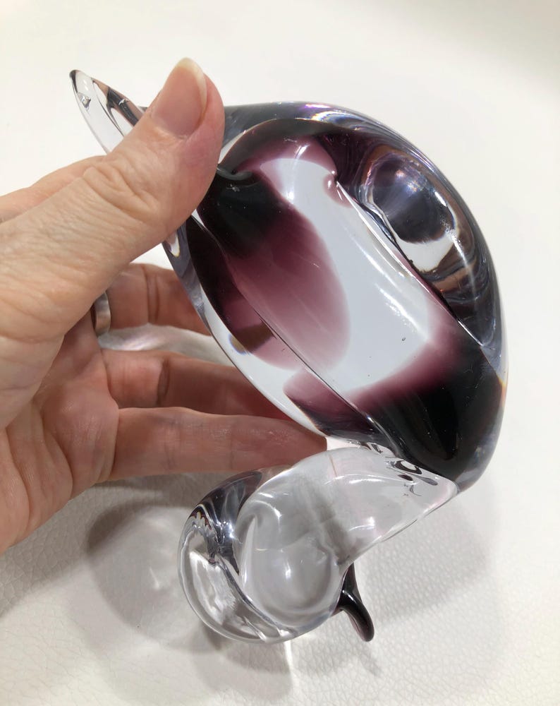 May include: A clear glass sculpture with a swirling design, featuring shades of purple and white. The art glass piece has a curved, organic shape and is held by a hand. The sculpture is a decorative item.