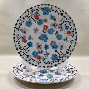 May include: Two white ceramic plates with floral designs. The larger plate features blue, red, and purple flowers with a black border. The smaller plate has a similar design. Both plates are displayed on a clear stand.