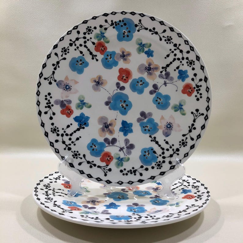 May include: Two white ceramic plates with floral designs. The larger plate features blue, red, and purple flowers with a black border. The smaller plate has a similar design. Both plates are displayed on a clear stand.