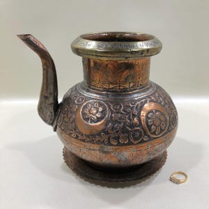 May include: An antique copper and silver-toned pitcher with an ornate floral and crescent moon design. The pitcher has a spout and a decorative rim. A small gold ring with clear stones sits next to the pitcher.
