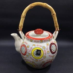 May include: A white ceramic teapot with a bamboo handle and a red lid. The teapot is decorated with a colorful floral pattern and the word "綠" in a black square.