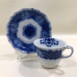 May include: A white porcelain teacup and matching saucer set featuring a deep blue transferware pattern. The saucer has a scalloped edge with intricate floral and lattice motifs, while the teacup displays a coordinating blue band and handle.