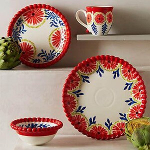 May include: A ceramic tableware set with a red, white, and blue floral pattern. The set includes a plate, a bowl, and a mug. The plate has a scalloped edge. The mug is on a shelf. Artichokes are on the surface.
