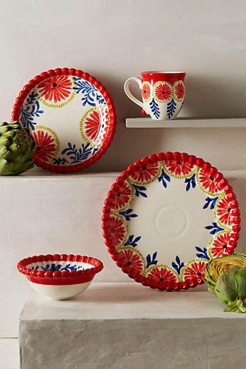 May include: A ceramic tableware set with a red, white, and blue floral pattern. The set includes a plate, a bowl, and a mug. The plate has a scalloped edge. The mug is on a shelf. Artichokes are on the surface.