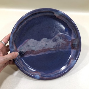 May include: A handmade ceramic plate with a deep indigo glaze. The plate features a unique design with shades of purple and white, creating a textured, artistic appearance. The rim has a slightly raised edge, adding to its handcrafted charm.