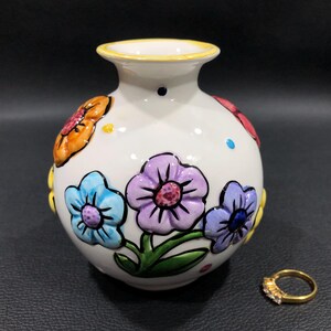 May include: A white ceramic vase with a yellow rim and colorful floral designs. The vase features blue, purple, and orange flowers. A gold ring with small stones is next to the vase.