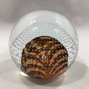 May include: A clear glass paperweight with a swirling brown and orange pattern inside. The paperweight is filled with small air bubbles.