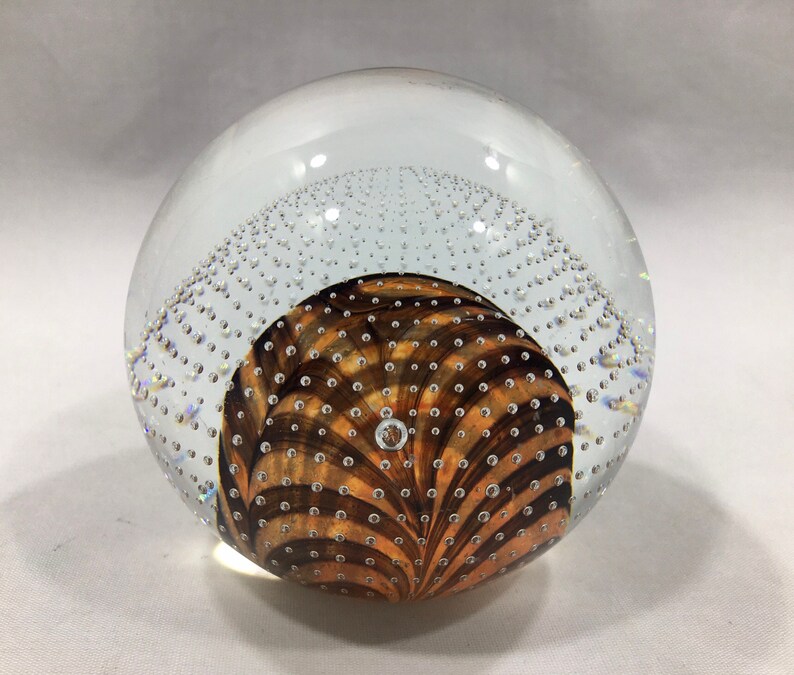 May include: A clear glass paperweight with a swirling brown and orange pattern inside. The paperweight is filled with small air bubbles.