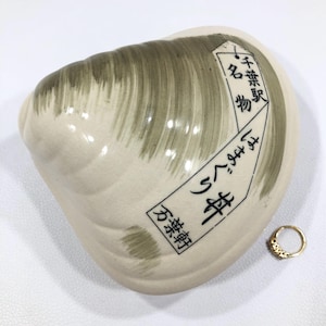 May include: A ceramic clam shell-shaped dish with a cream-colored base and olive green brushstrokes. The dish has Japanese text on a white label. A small gold ring with a decorative pattern is next to the dish.