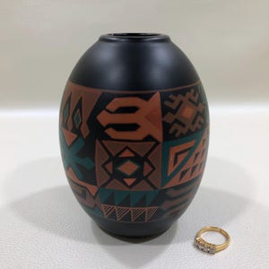 May include: Black ceramic vase with geometric patterns in shades of brown, teal, and orange. The vase has a rounded shape and a narrow opening. A gold ring with three small diamonds sits next to the vase.