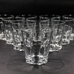 May include: A collection of clear, faceted drinking glasses. The glasses have a short, sturdy design with a geometric pattern. The image shows multiple glasses arranged in a group, suggesting a set or collection. The glasses are empty and appear to be made of glass.