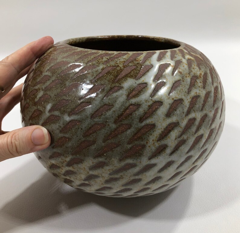 以下が含まれることがあります： 茶色と白の釉薬をかけた丸い陶器の花瓶。釉薬は斜めの線のテクスチャパターンになっています