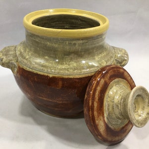 May include: A ceramic crock with a lid, featuring a brown and yellow glaze. The crock has a rounded body and a wide opening. The lid has a knob on top.