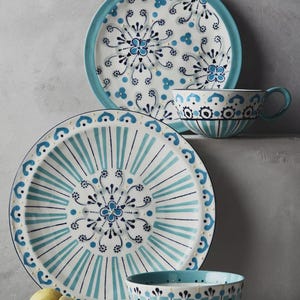 May include: A set of ceramic tableware featuring a white and blue floral design. The set includes a large plate, a smaller plate, a teacup, and a bowl. The plates have a turquoise border and the bowl has vertical stripes.