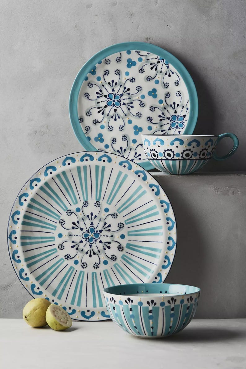 May include: A set of ceramic tableware featuring a white and blue floral design. The set includes a large plate, a smaller plate, a teacup, and a bowl. The plates have a turquoise border and the bowl has vertical stripes.