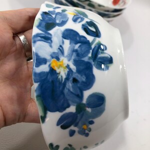 May include: White ceramic bowl with a blue floral design. The bowl features a large blue flower with yellow and green accents. Additional bowls with similar designs are visible in the background.
