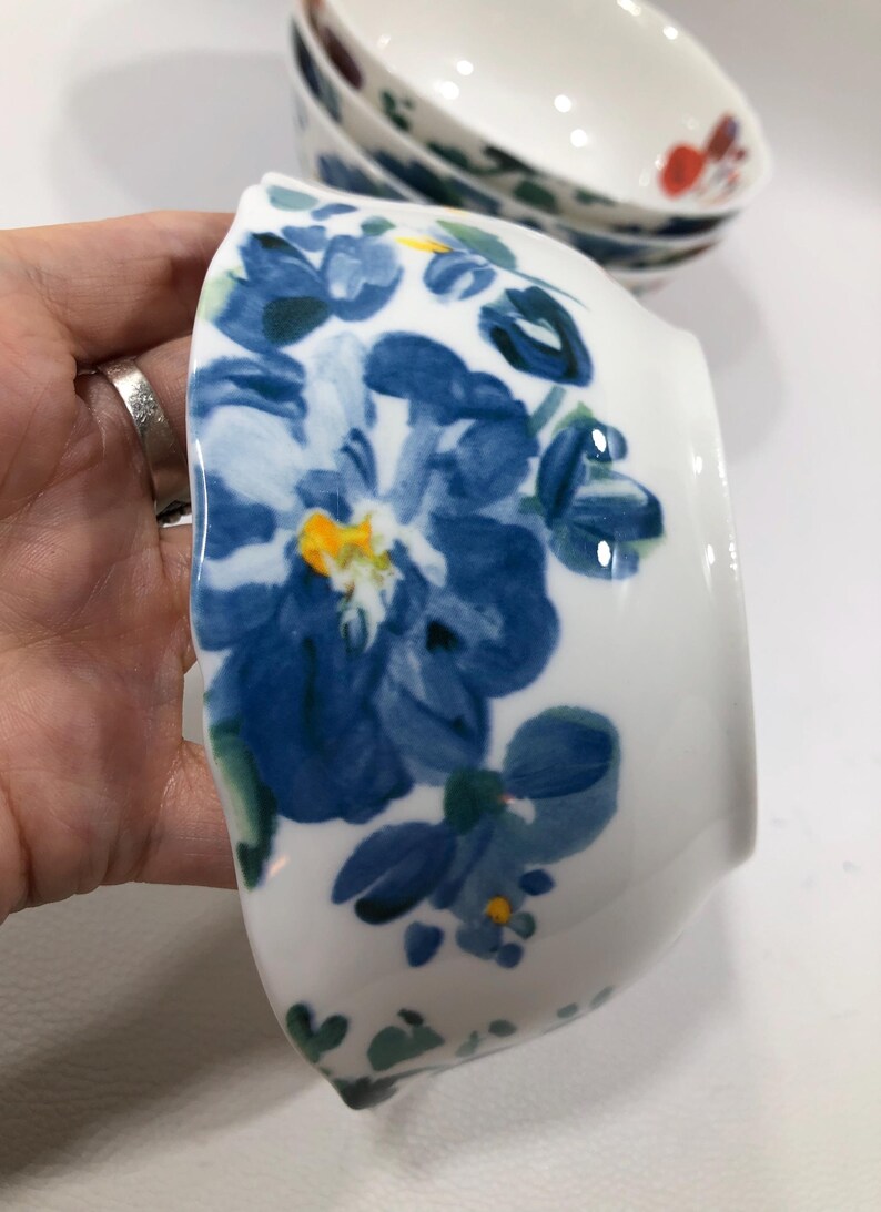 May include: White ceramic bowl with a blue floral design. The bowl features a large blue flower with yellow and green accents. Additional bowls with similar designs are visible in the background.
