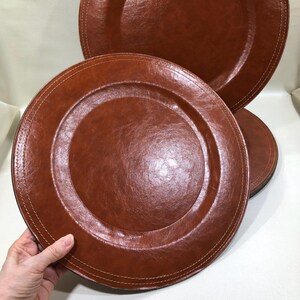 May include: A set of four brown leather-look charger plates. Each plate has a stitched edge and a raised center. The plates are a warm, reddish-brown color and appear to be made of a durable material. The plates are round.