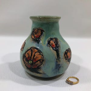 May include: A small, bulbous ceramic vase with a glossy light blue-green glaze, decorated with hand-painted orange and black floral motifs. A gold-toned ring with small clear stones is positioned to the right of the vase for scale reference.