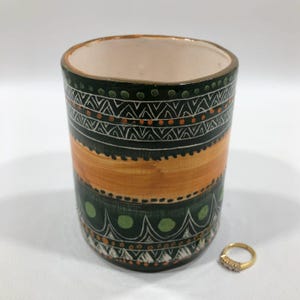 May include: A cylindrical ceramic vase with a cream interior, featuring a dark green and orange geometric pattern. The design includes white and orange accents. A gold ring with small stones sits beside the vase.