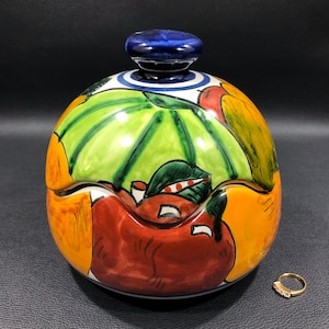 May include: A round ceramic trinket box with a lid, painted with colorful fruit designs. The box is decorated with yellow, orange, red, green, and blue paint. The lid has a blue knob.