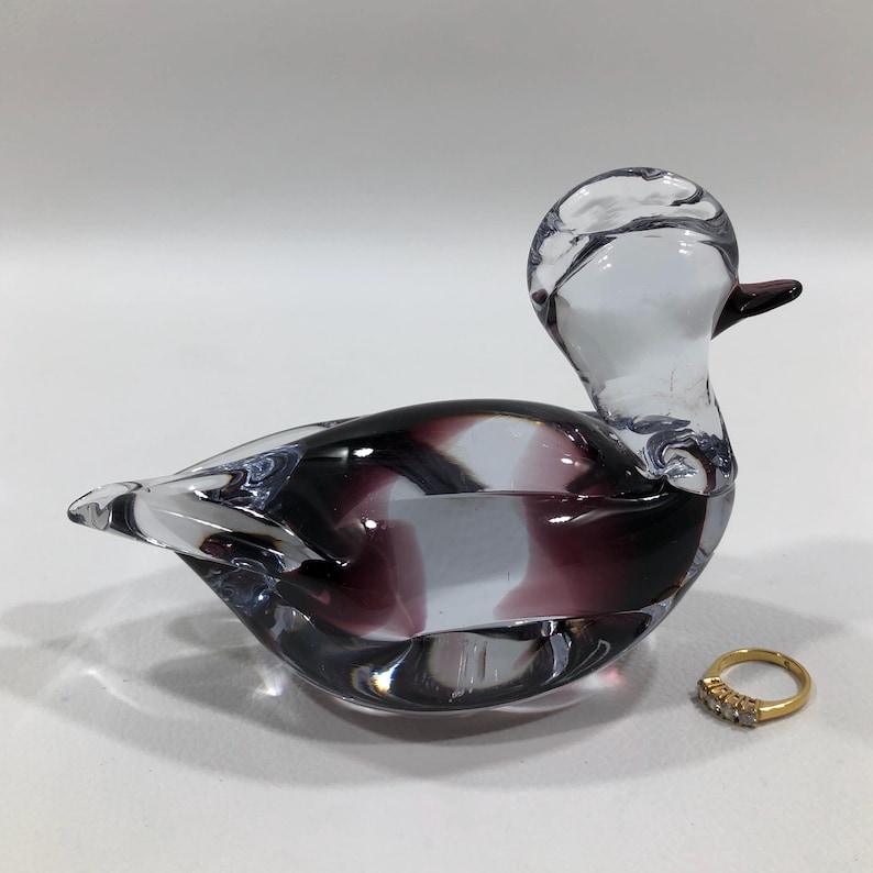 May include: A clear glass duck figurine with a purple and white design. The duck has a long neck and a small beak. A gold ring with three small diamonds sits next to the duck.