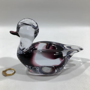 May include: A clear glass duck figurine with a purple and white swirl design. The duck has a small, dark purple beak and a gold ring with small stones is next to it.