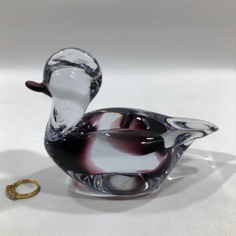 May include: A clear glass duck figurine with a purple and white swirl design. The duck has a small, dark purple beak and a gold ring with small stones is next to it.