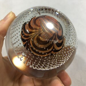 May include: A clear glass paperweight with a swirling pattern of brown and orange inside. The glass is filled with small air bubbles.