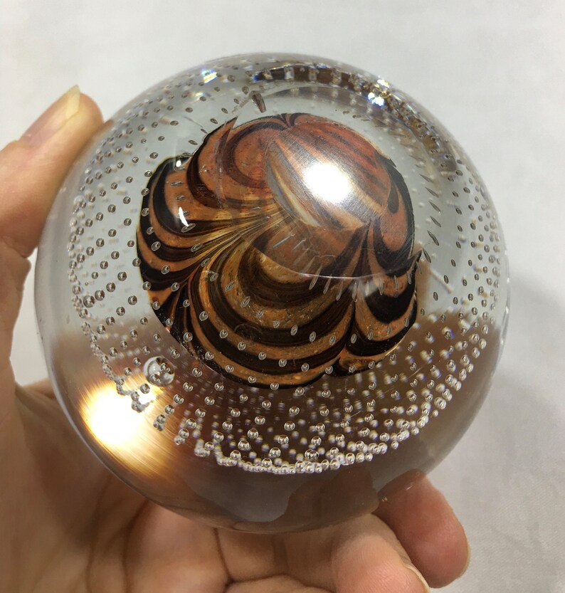 May include: A clear glass paperweight with a swirling pattern of brown and orange inside. The glass is filled with small air bubbles.