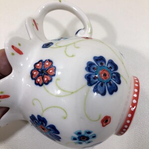Puede incluir: Taza de cer&aacute;mica blanca con asa, decorada con motivos florales azules y rojos y enredaderas verdes. El borde presenta una banda roja con puntos blancos. La taza est&aacute; sujeta por una mano.