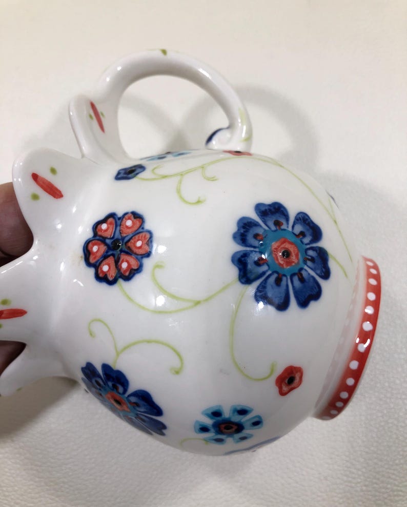 Puede incluir: Taza de cer&aacute;mica blanca con asa, decorada con motivos florales azules y rojos y enredaderas verdes. El borde presenta una banda roja con puntos blancos. La taza est&aacute; sujeta por una mano.