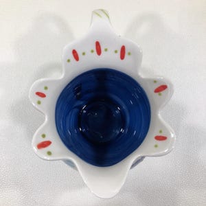 Puede incluir: Una crema de cer&aacute;mica con un borde blanco festoneado y un interior azul oscuro. El borde est&aacute; decorado con puntos rojos y verdes. La crema tiene un pico y un asa. El dise&ntilde;o recuerda a una flor.