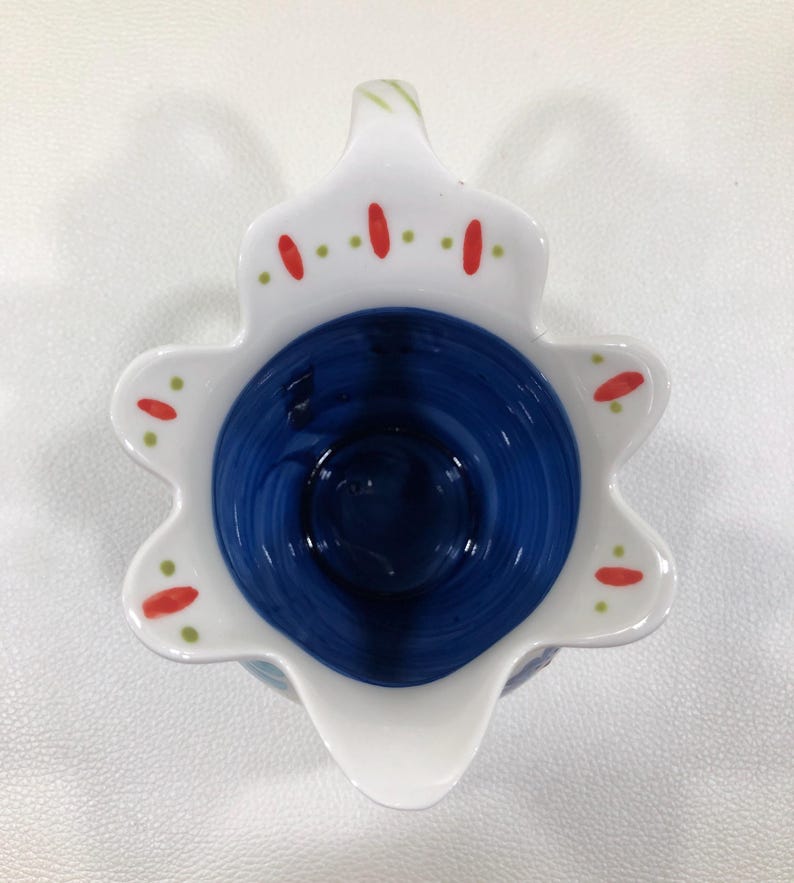 Puede incluir: Una crema de cer&aacute;mica con un borde blanco festoneado y un interior azul oscuro. El borde est&aacute; decorado con puntos rojos y verdes. La crema tiene un pico y un asa. El dise&ntilde;o recuerda a una flor.