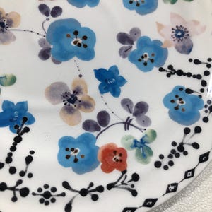 May include: White ceramic plate with a hand-painted floral design. The plate features blue, red, and purple flowers with green leaves. A black geometric border decorates the edge of the plate. The plate is a decorative item.