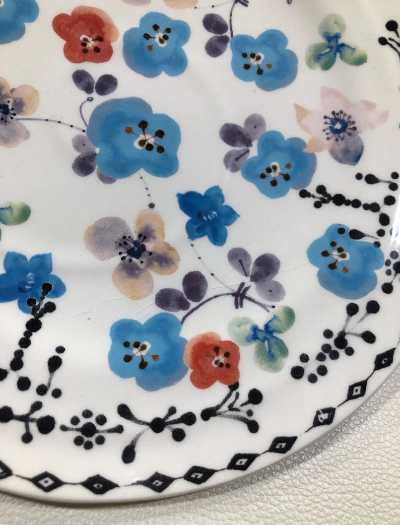May include: White ceramic plate with a hand-painted floral design. The plate features blue, red, and purple flowers with green leaves. A black geometric border decorates the edge of the plate. The plate is a decorative item.