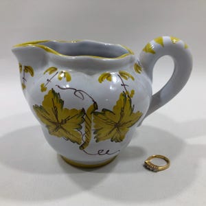 May include: White ceramic pitcher with a yellow rim and handle, decorated with yellow and green grape leaf designs. A gold ring with three small diamonds sits next to the pitcher.
