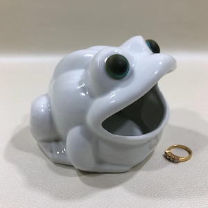 May include: A white ceramic frog-shaped container with large, expressive eyes. The frog has an open mouth, revealing an interior space. A gold ring with small stones sits beside the frog. The frog is a decorative item.