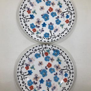 May include: Two white ceramic plates with a floral design. The plates feature blue, red, and purple flowers with a black dotted border. The flowers are scattered across the surface of the plates.
