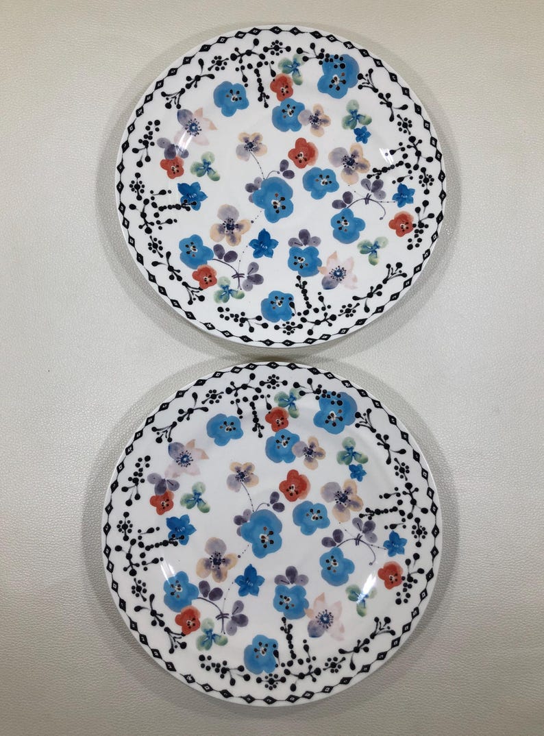 May include: Two white ceramic plates with a floral design. The plates feature blue, red, and purple flowers with a black dotted border. The flowers are scattered across the surface of the plates.