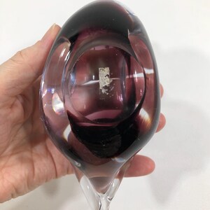 May include: A hand is holding a clear glass vase with a deep purple interior and a clear, pointed base. The vase has a rounded top and a smooth, polished surface. The color transitions from clear to deep purple.